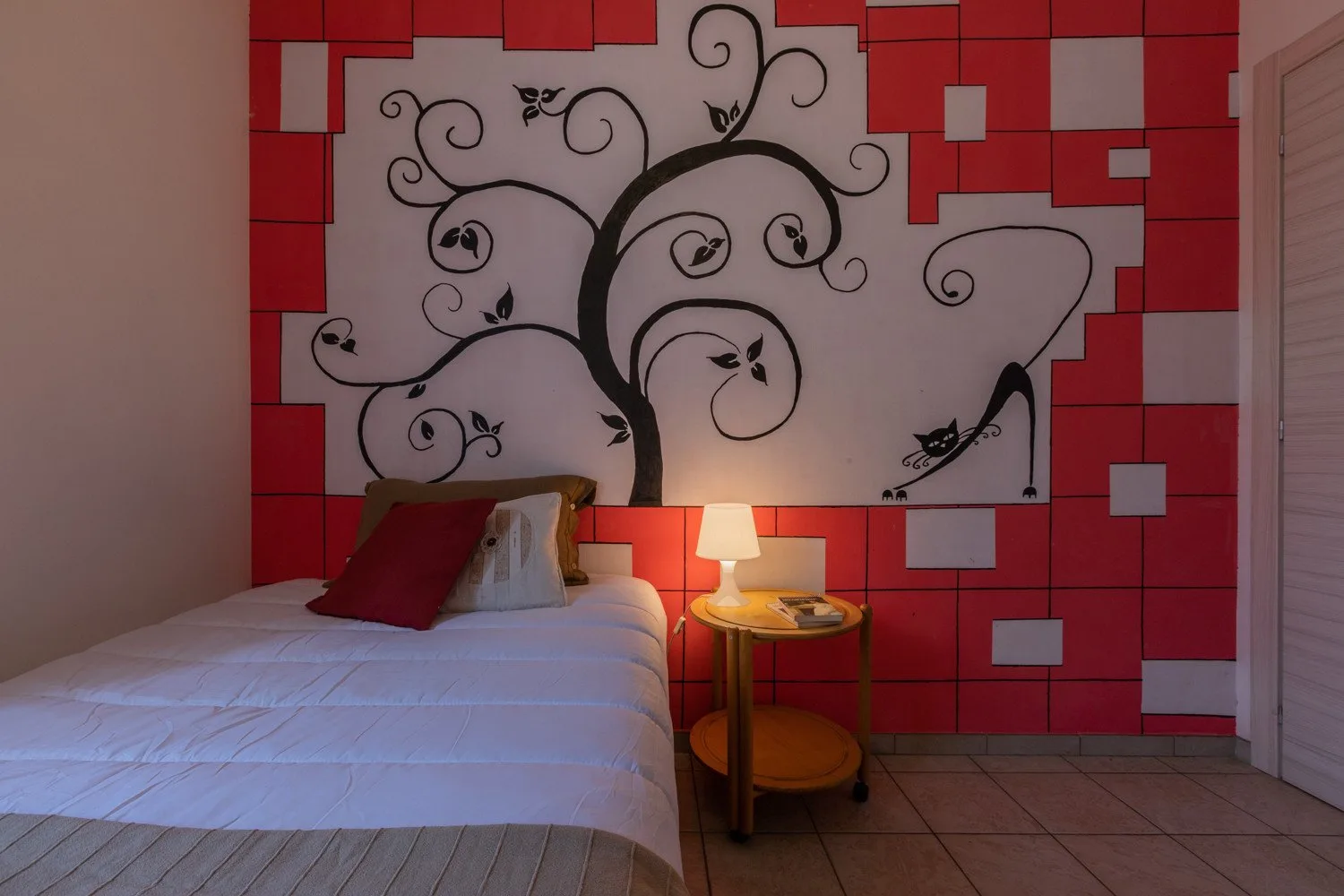 A bedroom with a bed beside a red wall featuring a large black tree mural.