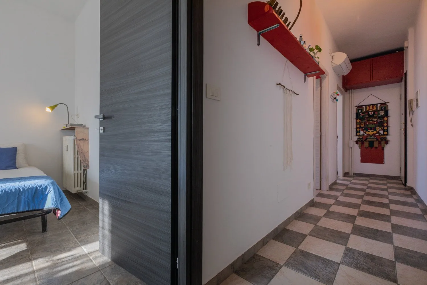 Un pasillo con suelo de baldosas y un estante rojo conduce a un dormitorio con una cama.