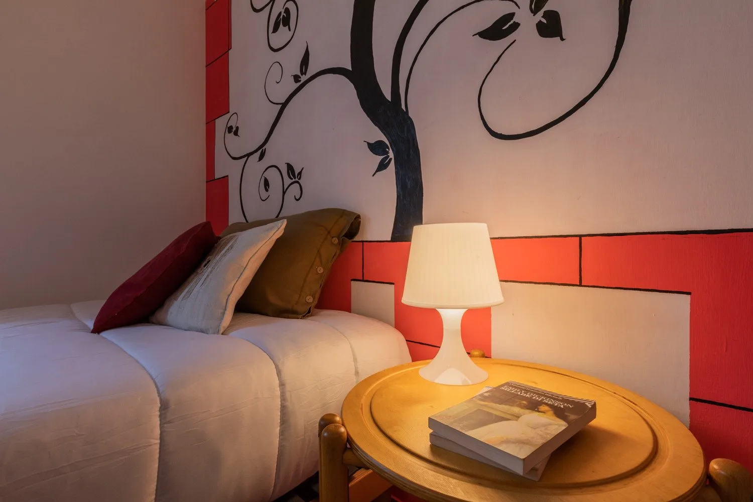 A bedroom with a bed, a bedside table, and a lamp beneath a painted wall mural.