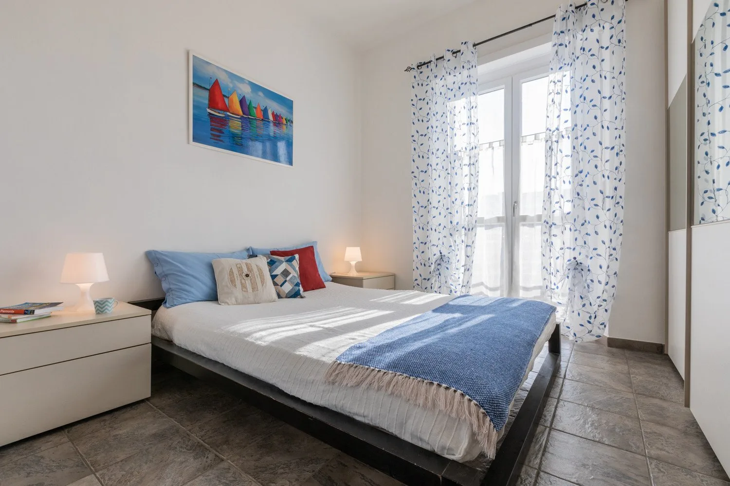 A bright bedroom with a bed, blue bedding, and patterned curtains.