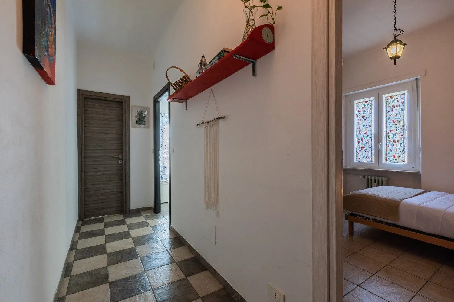 Un pasillo con suelo de baldosas da a un dormitorio con una cama y ventanas estampadas.