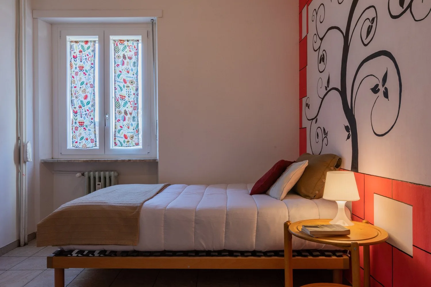 A bedroom with a neatly made bed, a bedside lamp, and a colorful window.