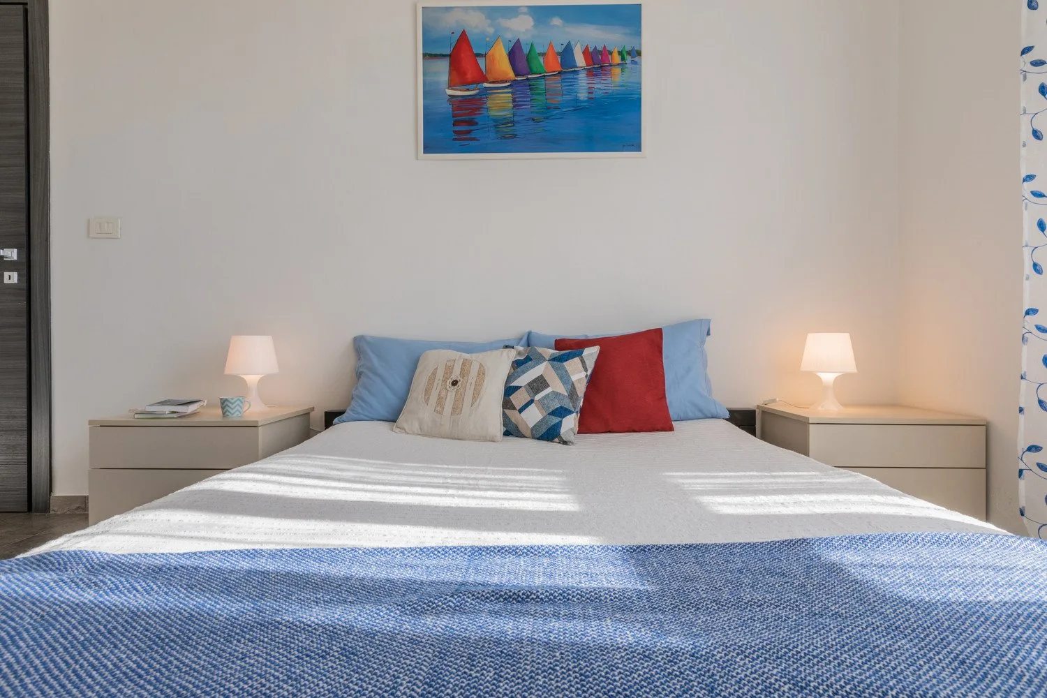 Un dormitorio con una cama tendida, mesitas de noche y una colorida pintura de veleros.
