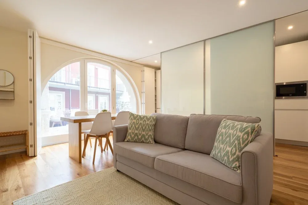 A bright living area with a gray sofa beside a dining table and arched window.