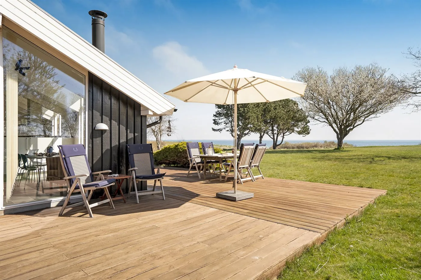 Una terraza de madera junto a una casa con paredes de vidrio tiene asientos al aire libre y una sombrilla.