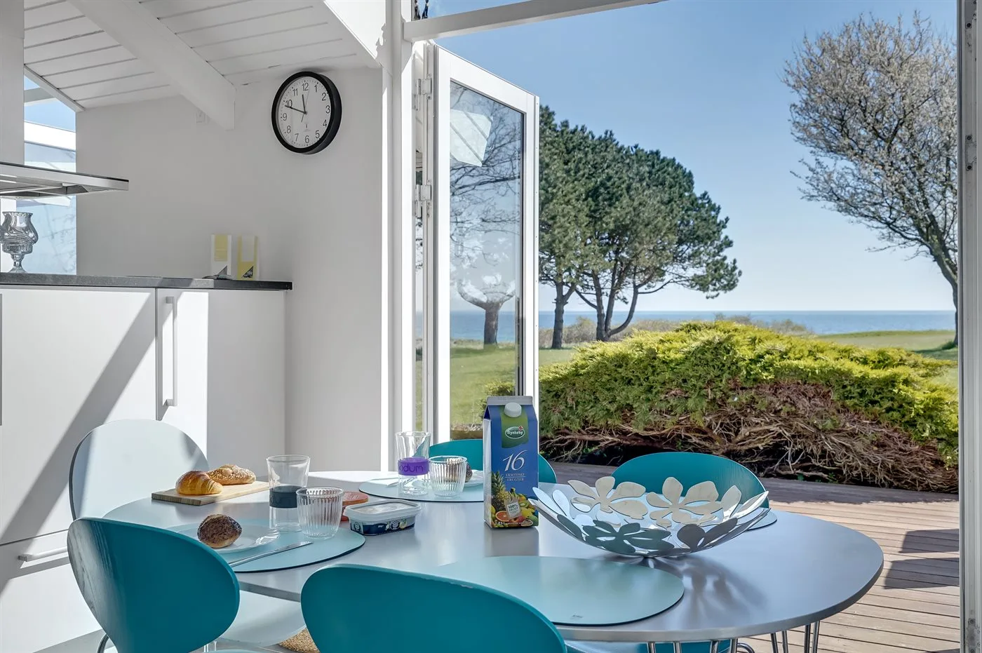 Comedor con mesa de cristal junto a puertas abiertas y vistas al mar.