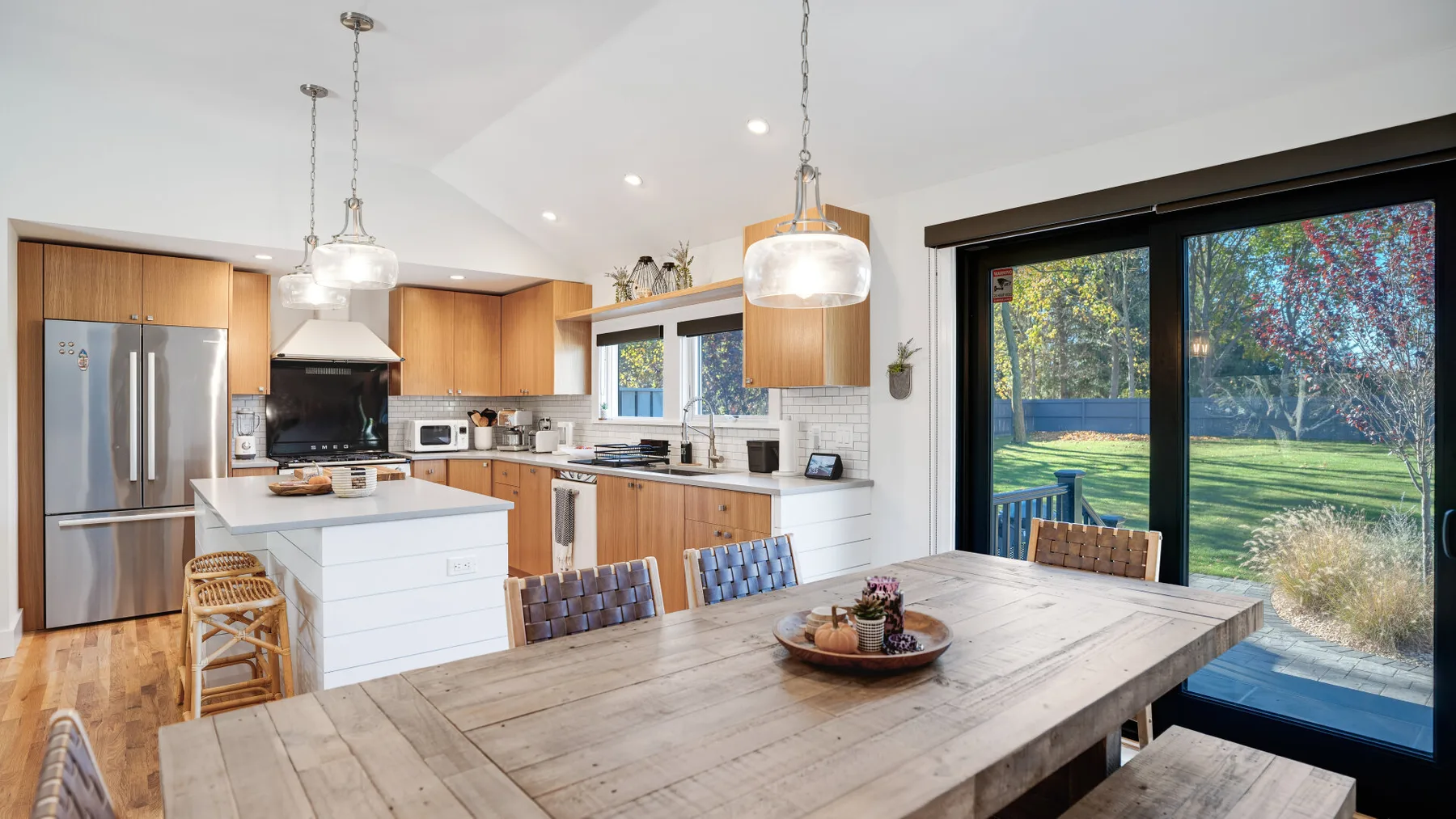 Una cocina abierta con comedor tiene una mesa grande de madera junto a puertas de vidrio.