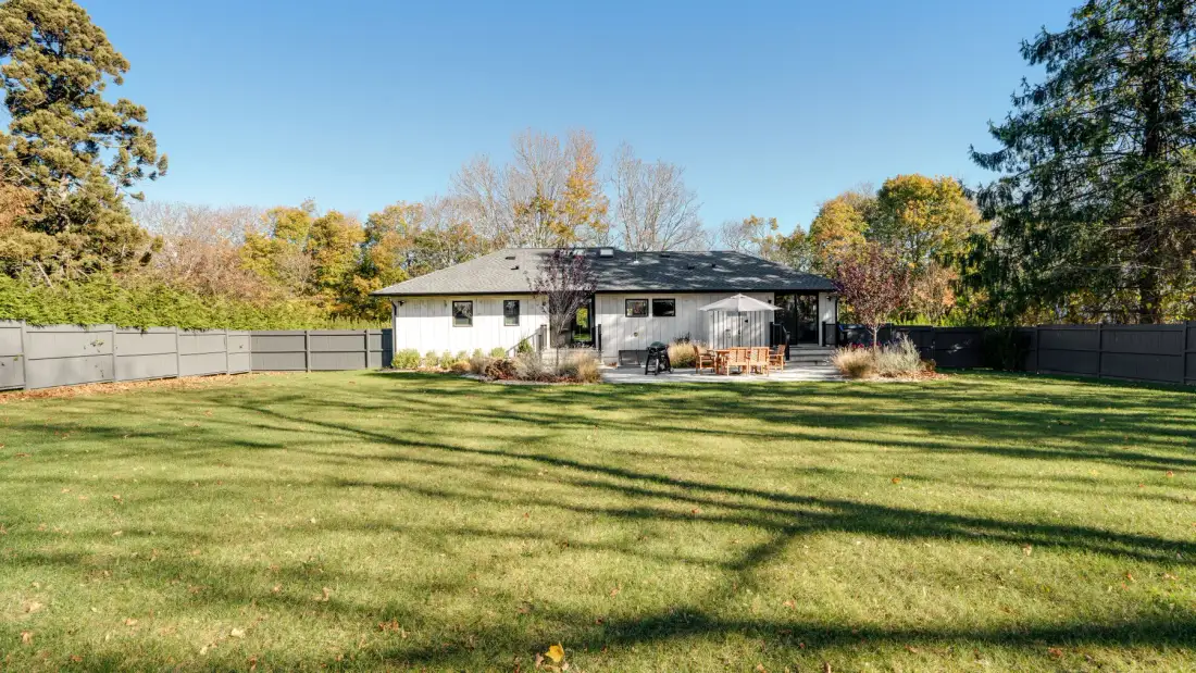 A single-story house sits beyond a broad grassy backyard bordered by trees and fencing.