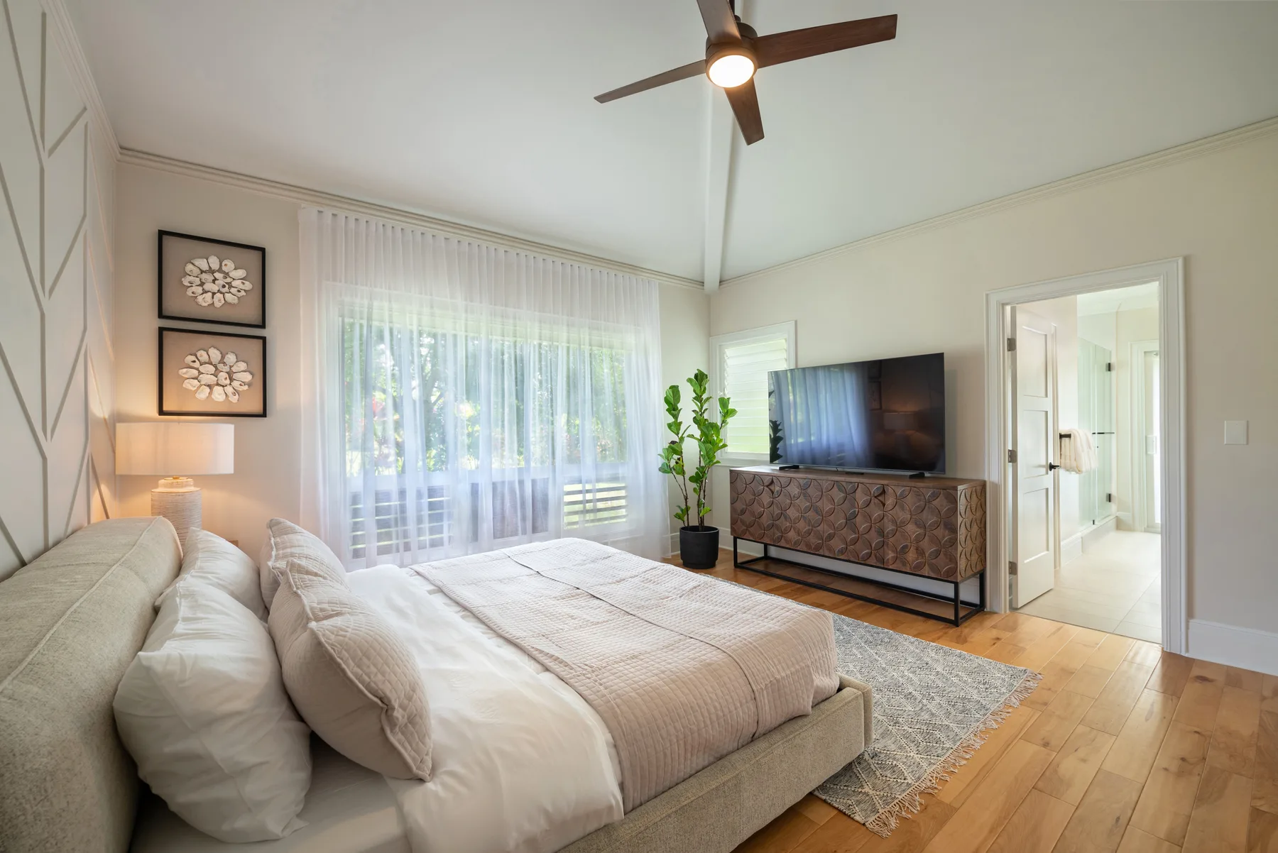 Un dormitorio luminoso con una cama tendida, una gran ventana con cortinas y un televisor.