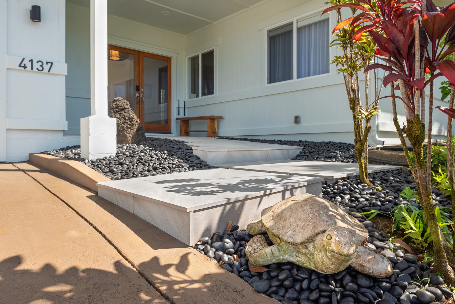 La entrada de una casa da a un camino pavimentado bordeado de piedras oscuras y plantas tropicales.