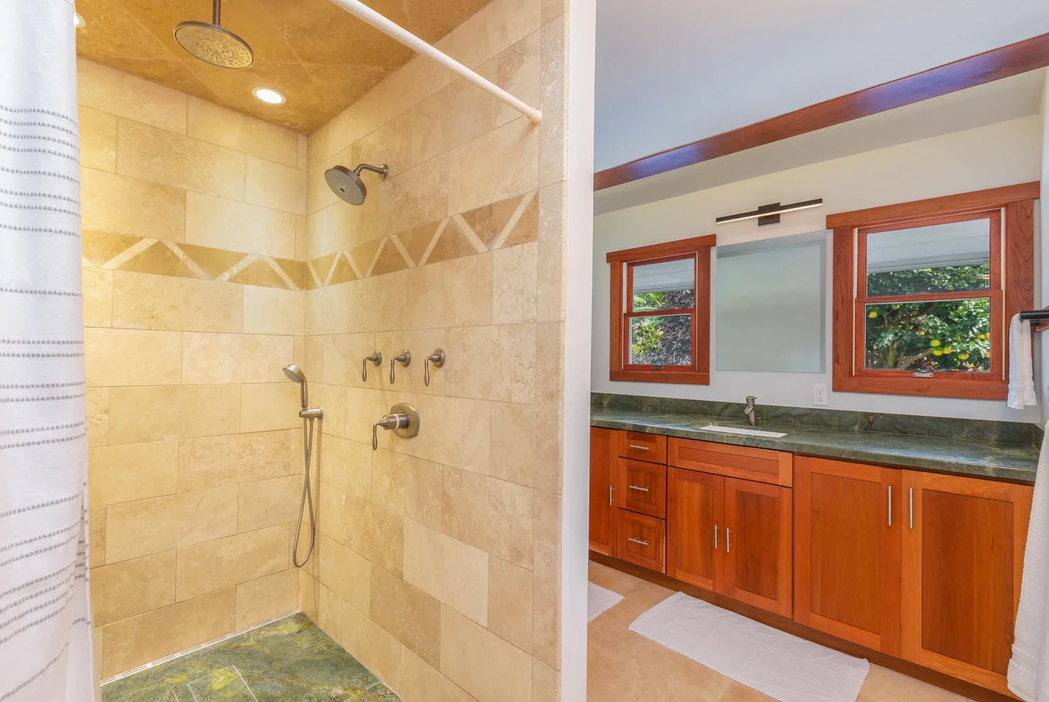 A tiled shower beside a bathroom vanity with wooden cabinets and windows.