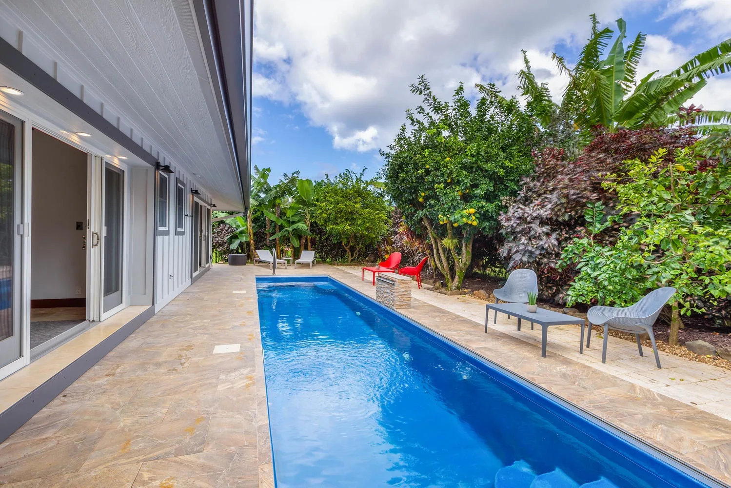 An outdoor swimming pool beside a paved patio, with lounge seating and tropical greenery.