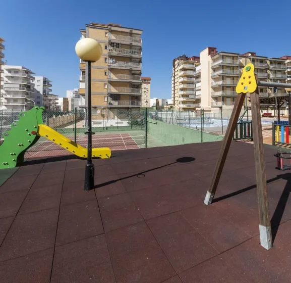 An outdoor playground with climbing equipment stands beside fenced sports courts and apartment buildings.