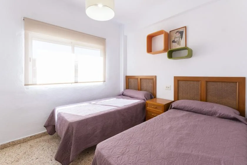 A bright bedroom with two beds, a window, and colorful wall shelves.