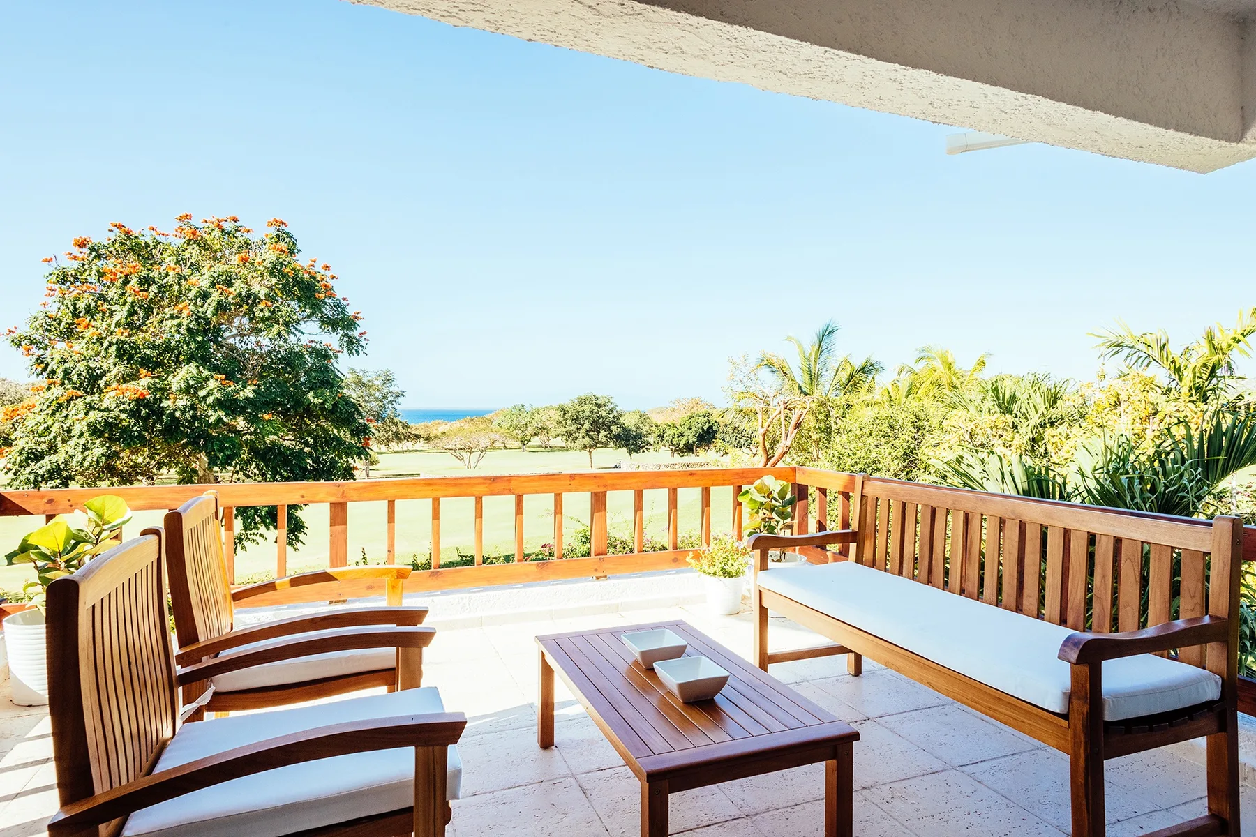 An outdoor patio has wooden seating and a table overlooking palm trees and the sea.