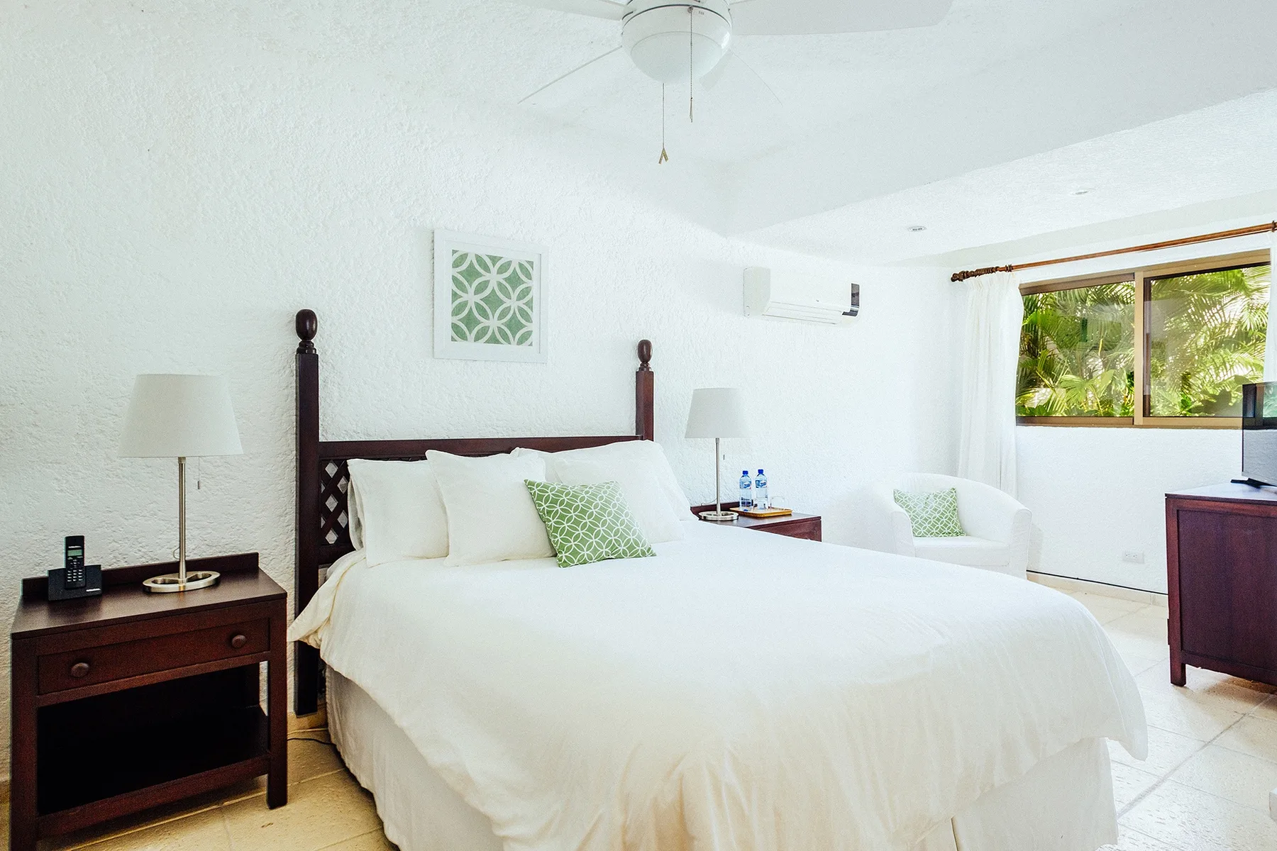 A bright bedroom with a large bed, bedside tables, and a window overlooking greenery.