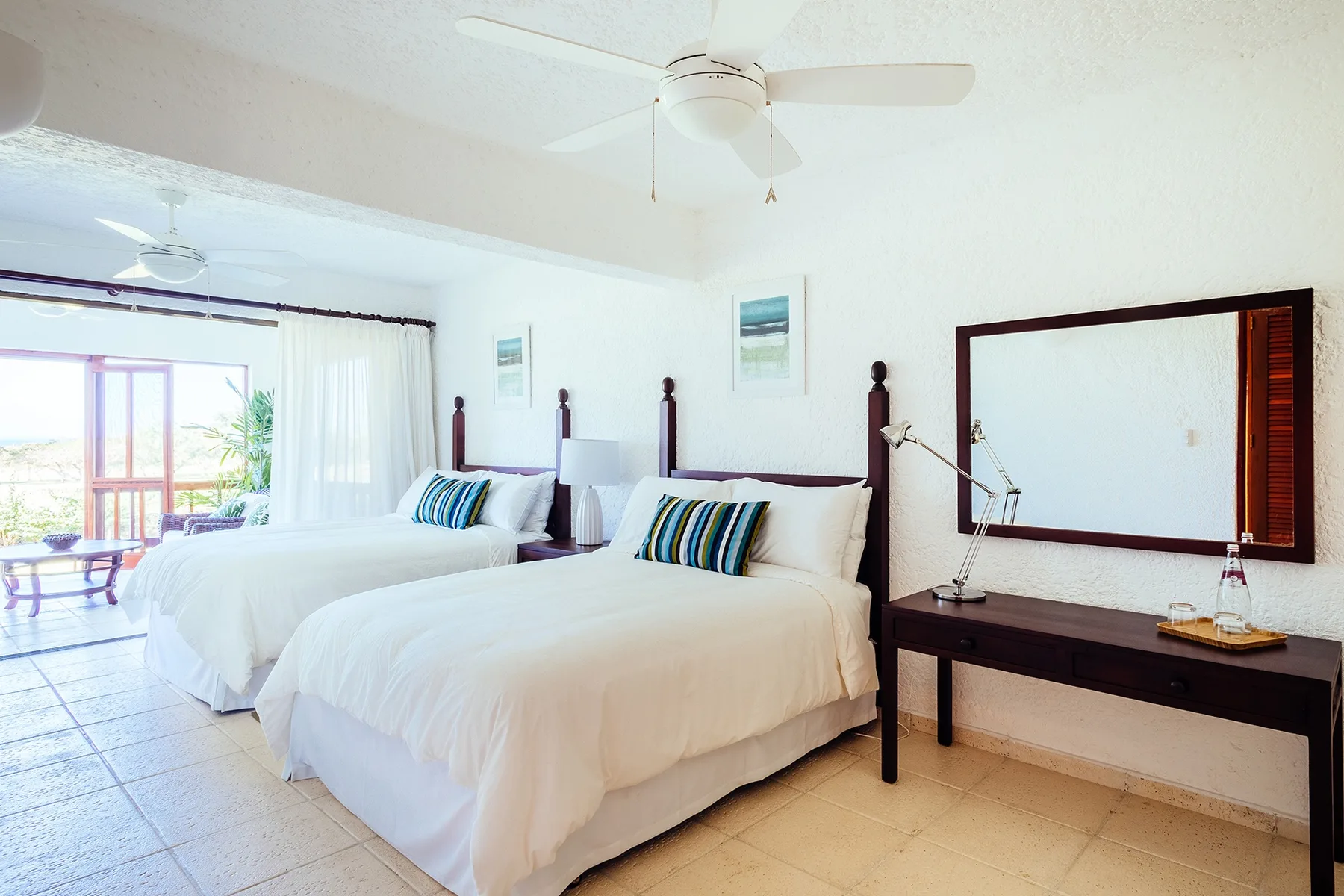 A bright bedroom with two beds dressed in white linens and a desk beneath a mirror.