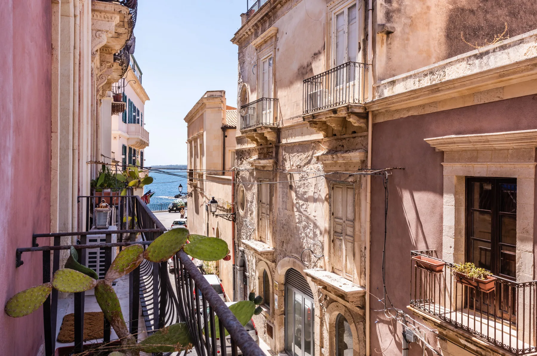 Una calle estrecha entre edificios antiguos se abre al mar, con balcones y plantas.