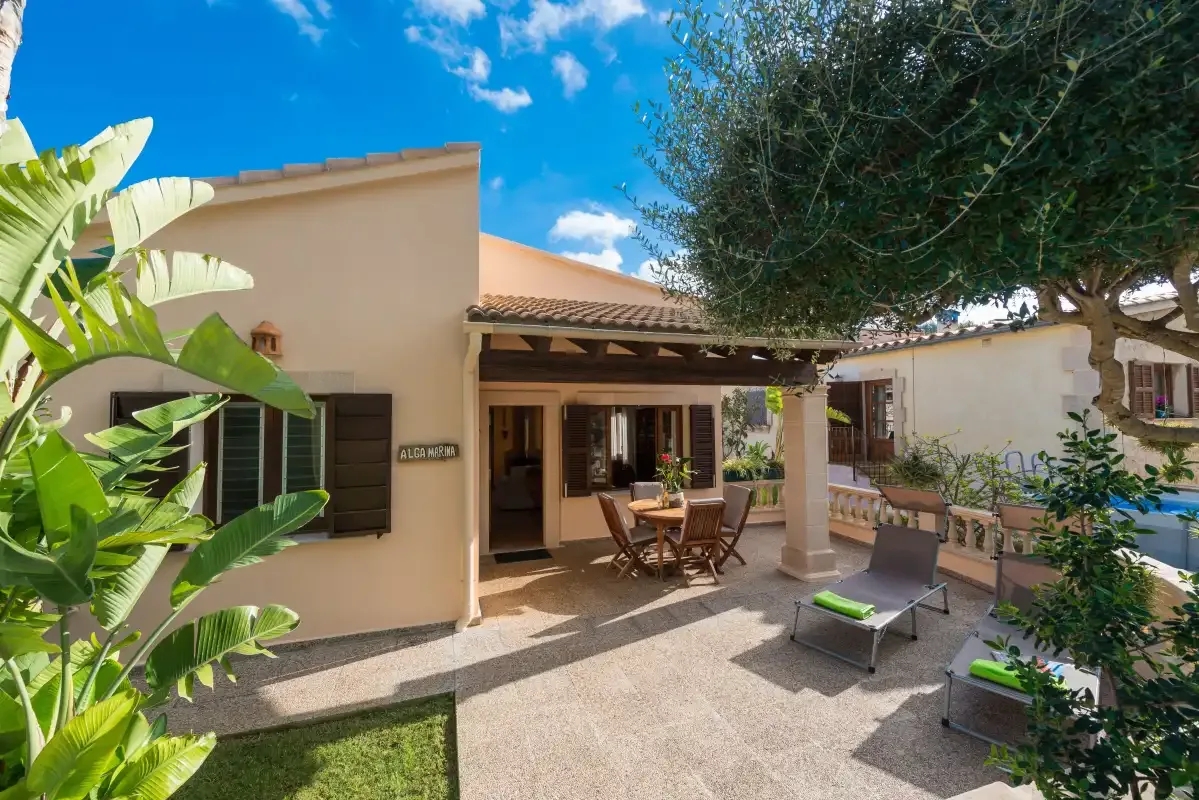 A house opens onto a sunny patio with outdoor seating and leafy plants.
