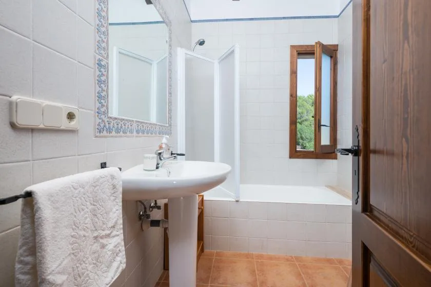 A tiled bathroom with a sink, mirror, and bathtub beneath an open window.