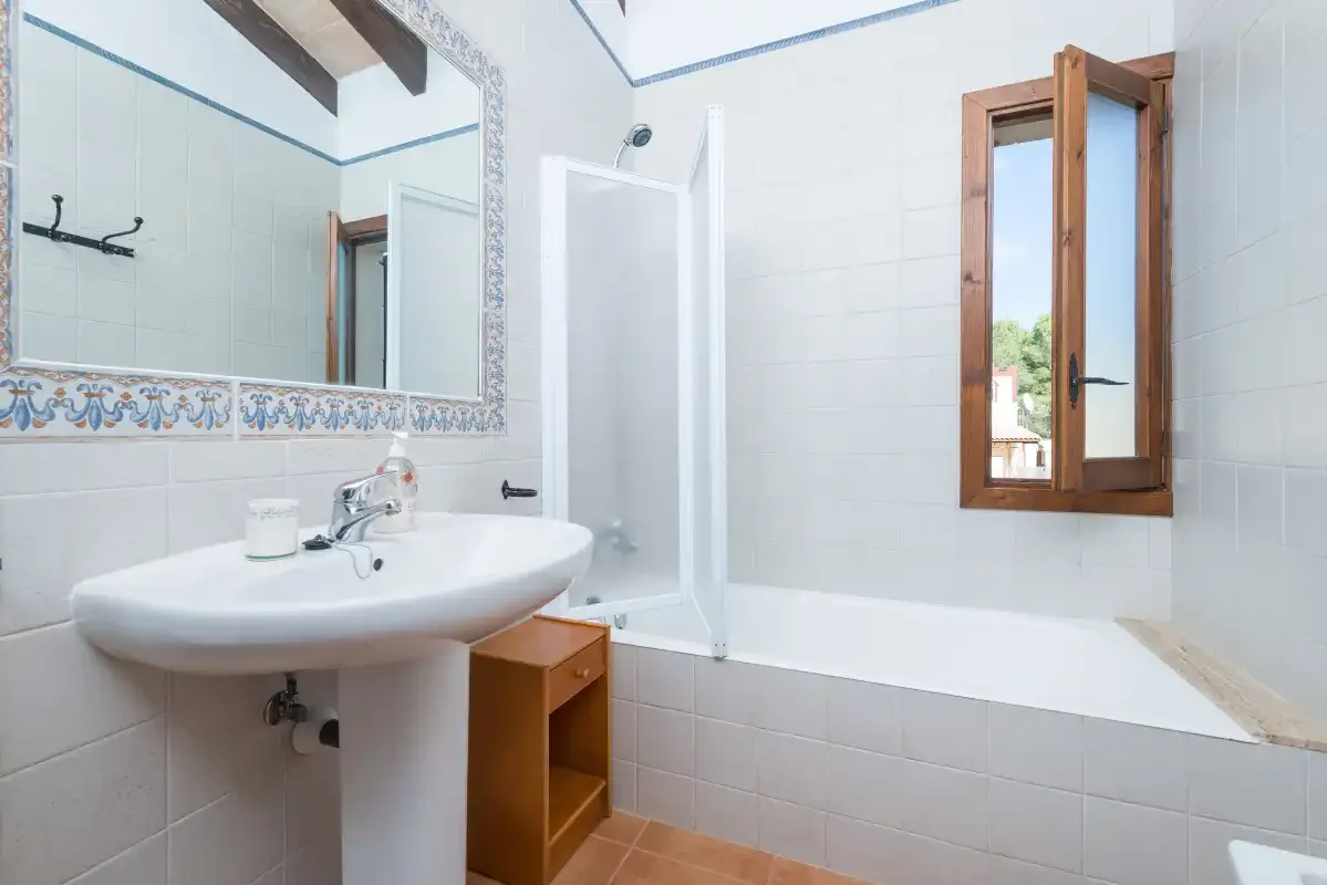 A tiled bathroom with a sink, bathtub, and open wooden window.