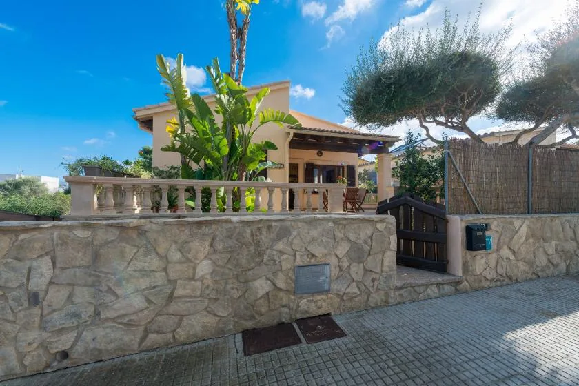 A house stands behind a stone boundary wall, with a gate and leafy plants.