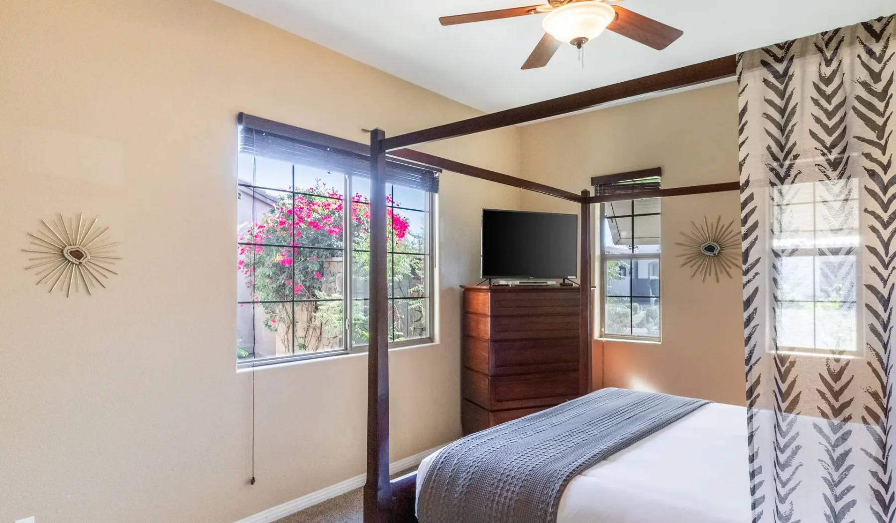 A bedroom with a four-poster bed, television, and windows overlooking flowering plants.