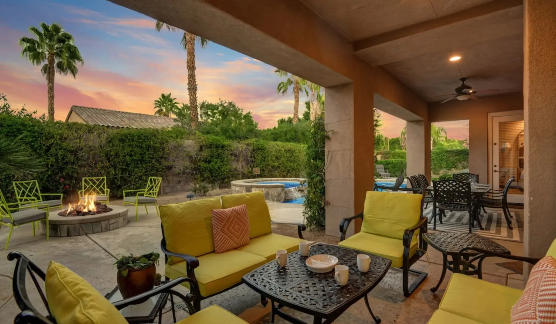 A covered patio has outdoor seating, a fire pit, and a pool in the background.