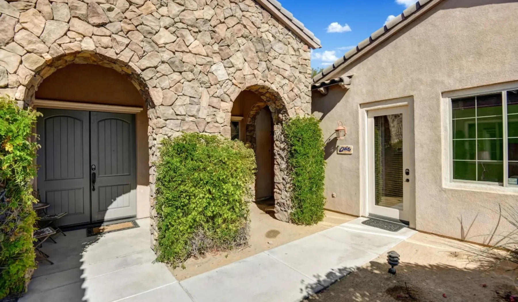A stone-fronted house entrance opens onto a sunlit paved walkway.