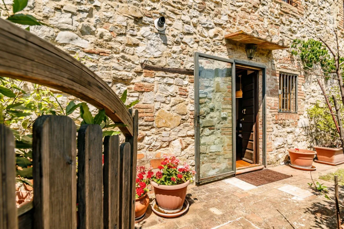 A stone building entrance opens onto a sunlit patio with potted flowers.