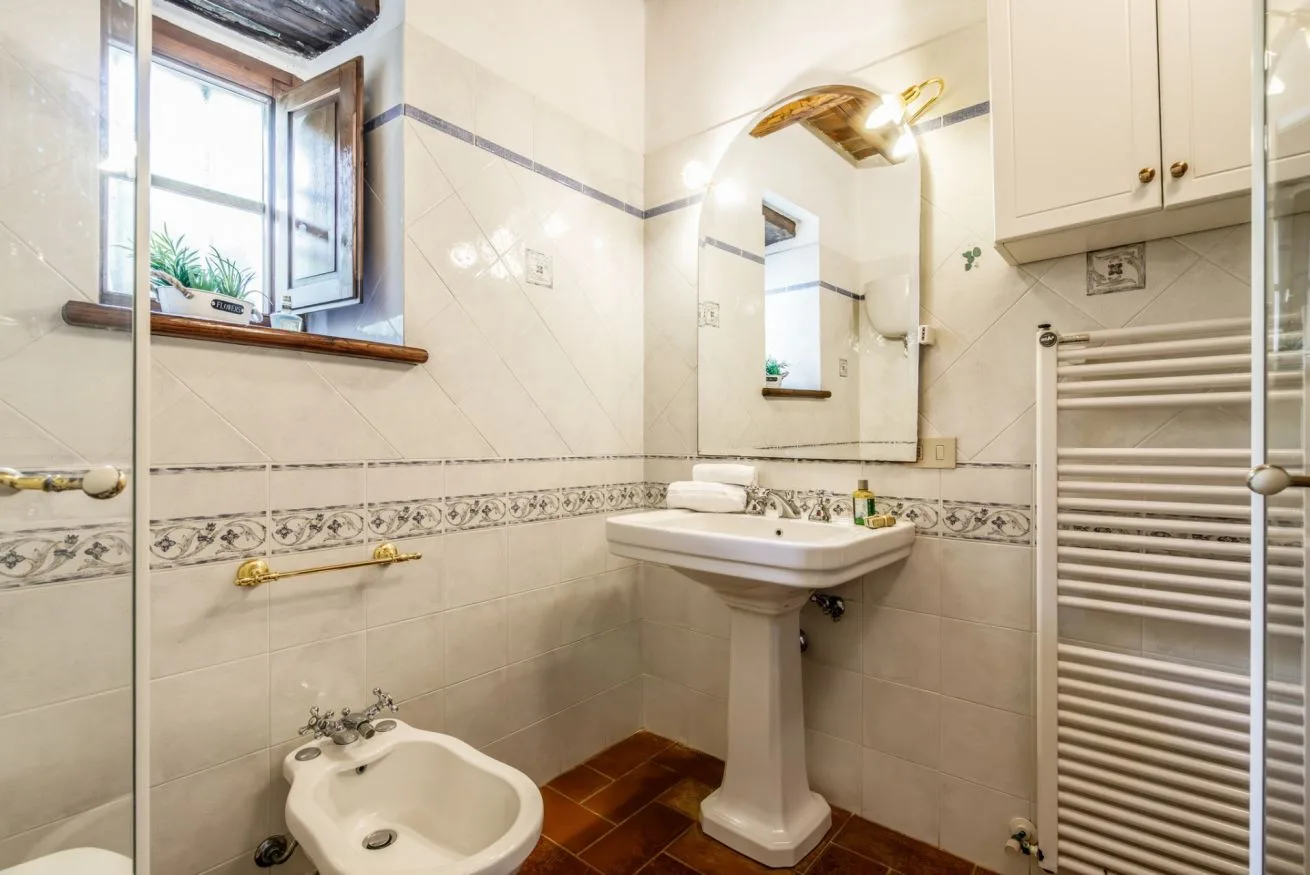 A tiled bathroom with a pedestal sink, mirror, and window.