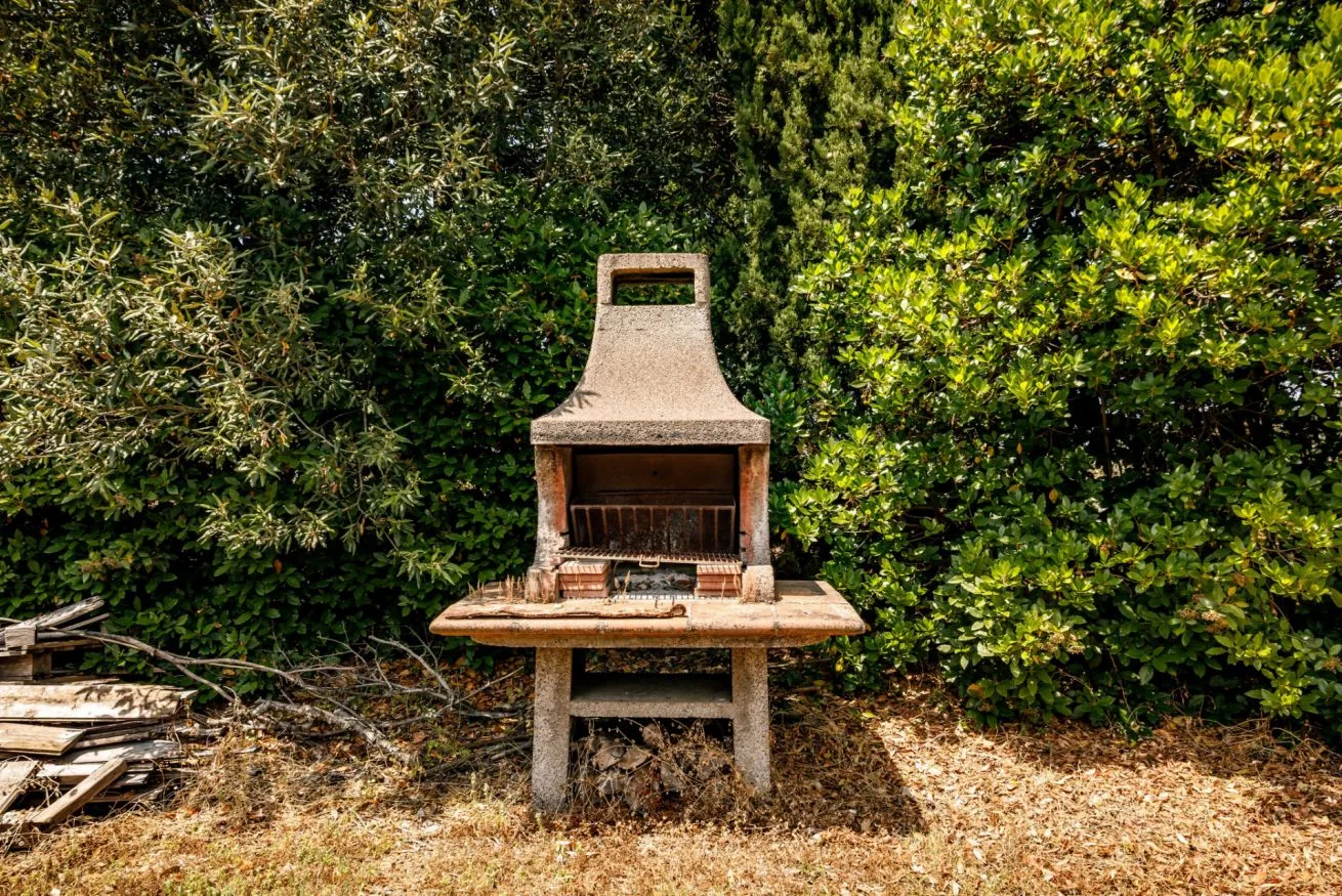 An outdoor masonry barbecue grill stands in front of dense greenery.