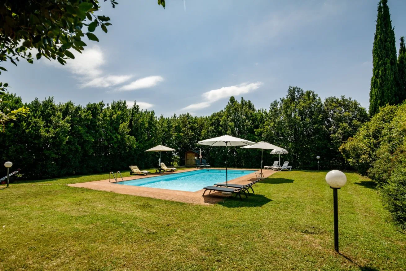 An outdoor swimming pool sits in a grassy garden with trees and umbrellas.