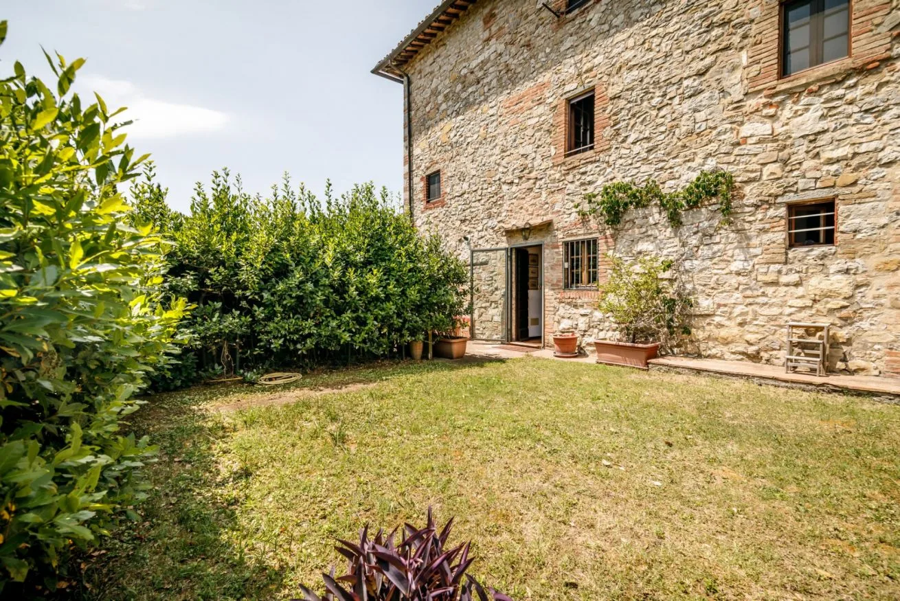 A grassy garden sits beside a stone house, with shrubs along the wall.