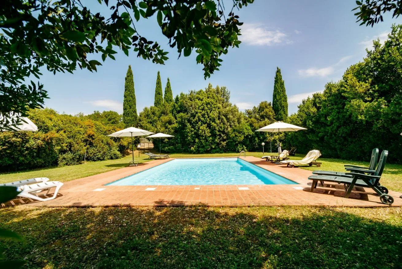 An outdoor swimming pool is surrounded by lawn, trees, and lounge seating.