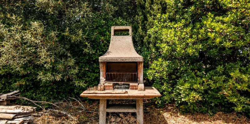 An outdoor masonry barbecue stands in front of dense greenery.