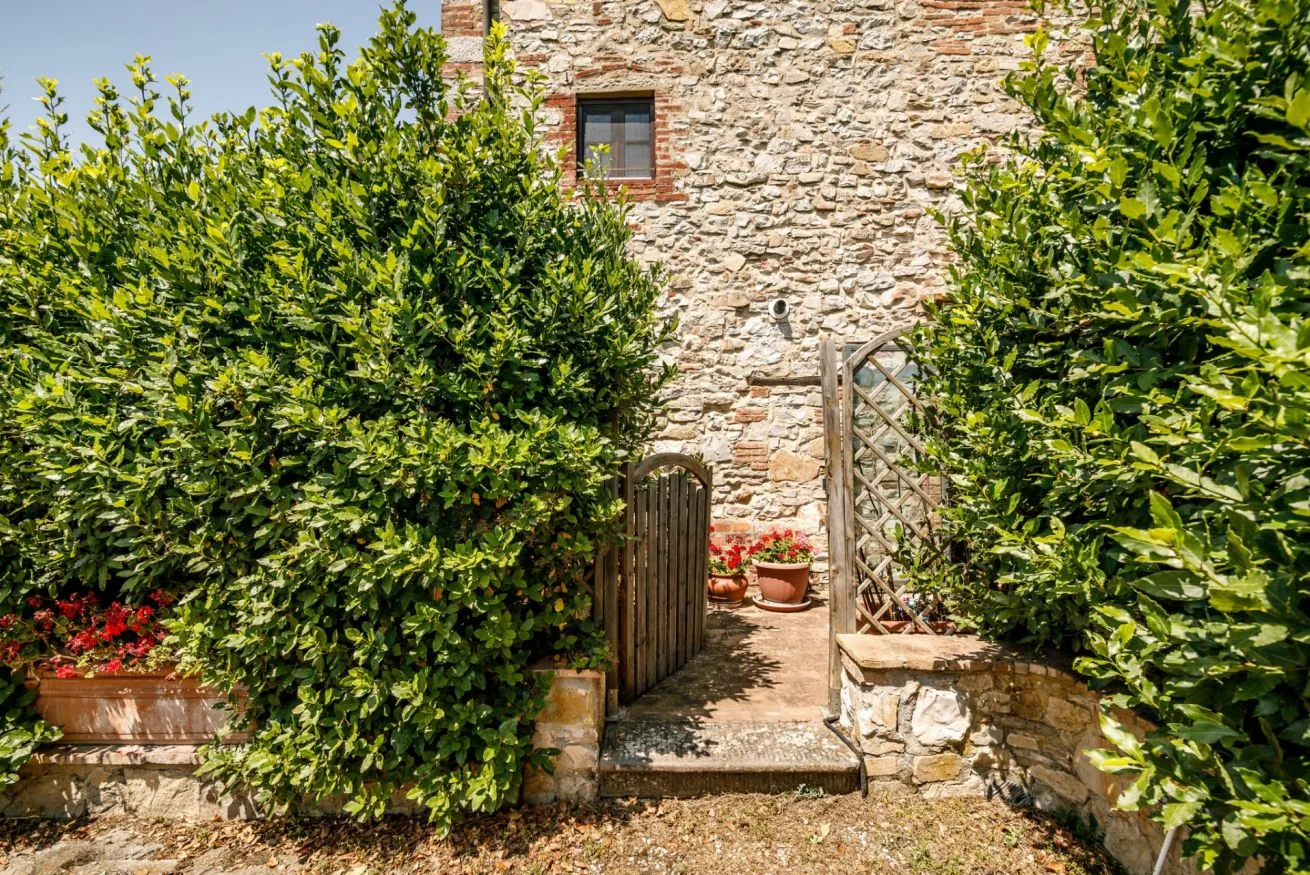 A stone building entrance is framed by leafy shrubs and potted flowers.