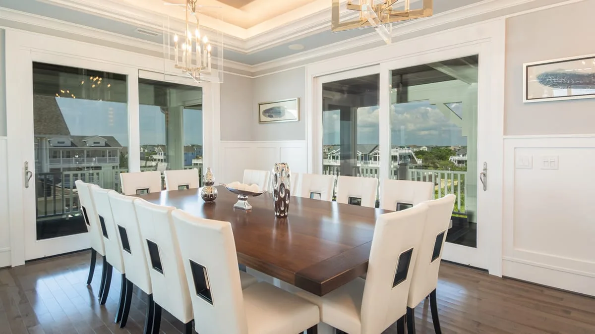 A bright dining room with a long wooden table surrounded by white seating.
