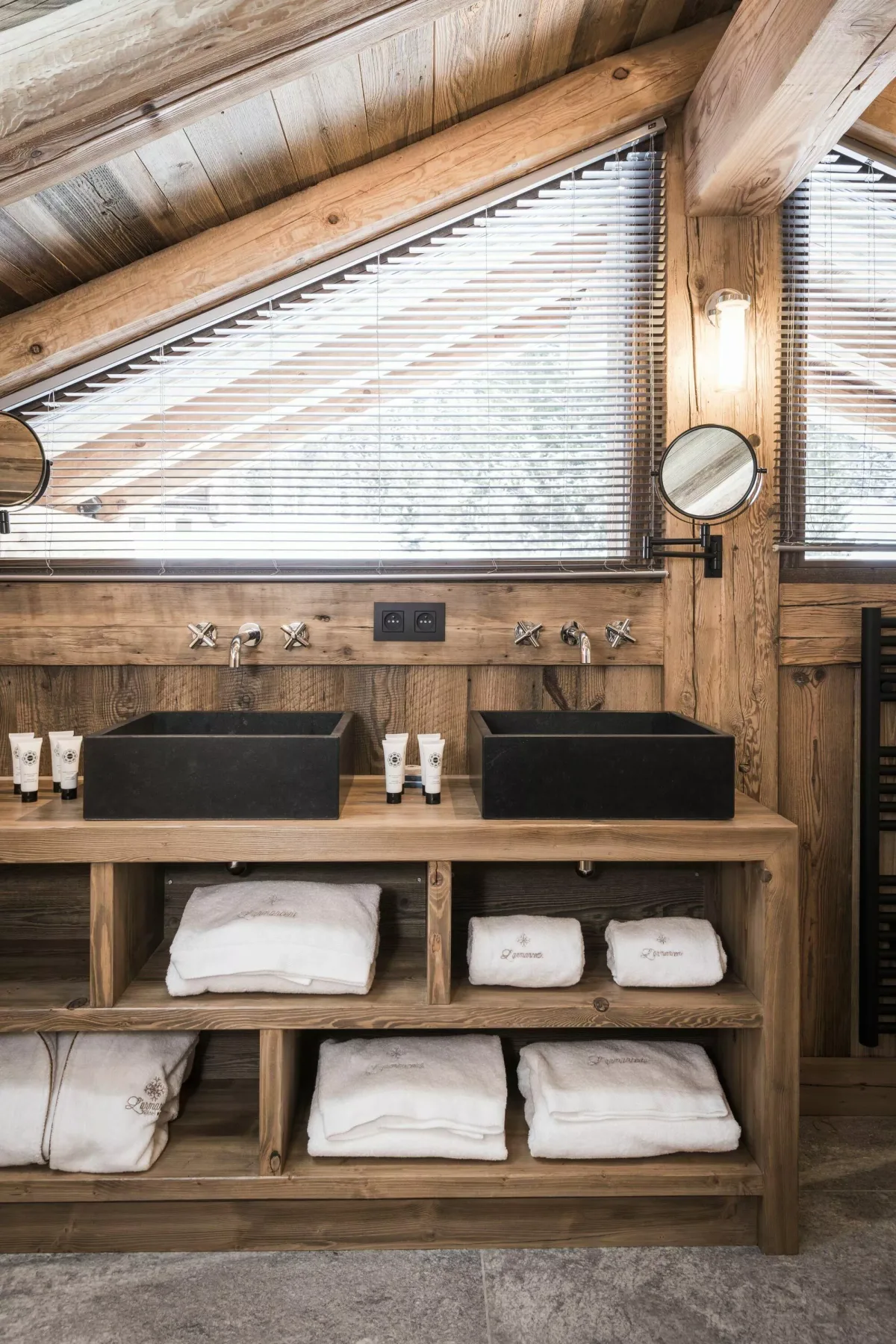 Un tocador de baño rústico con dos lavabos negros está bajo ventanas inclinadas.