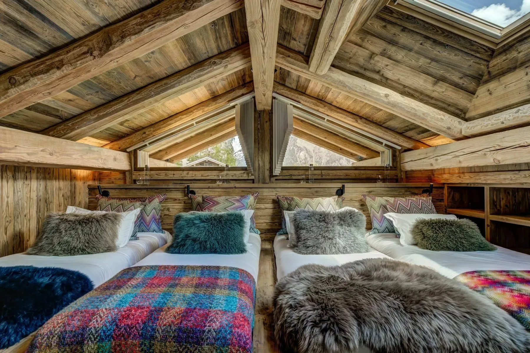 Un dormitorio rústico con camas bajo un techo de madera inclinado y ventanas triangulares.