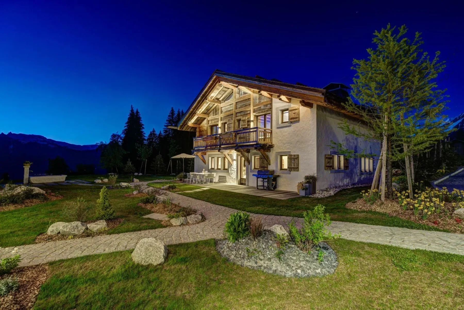 Una casa de estilo alpino iluminada se alza junto a un jardín al anochecer.