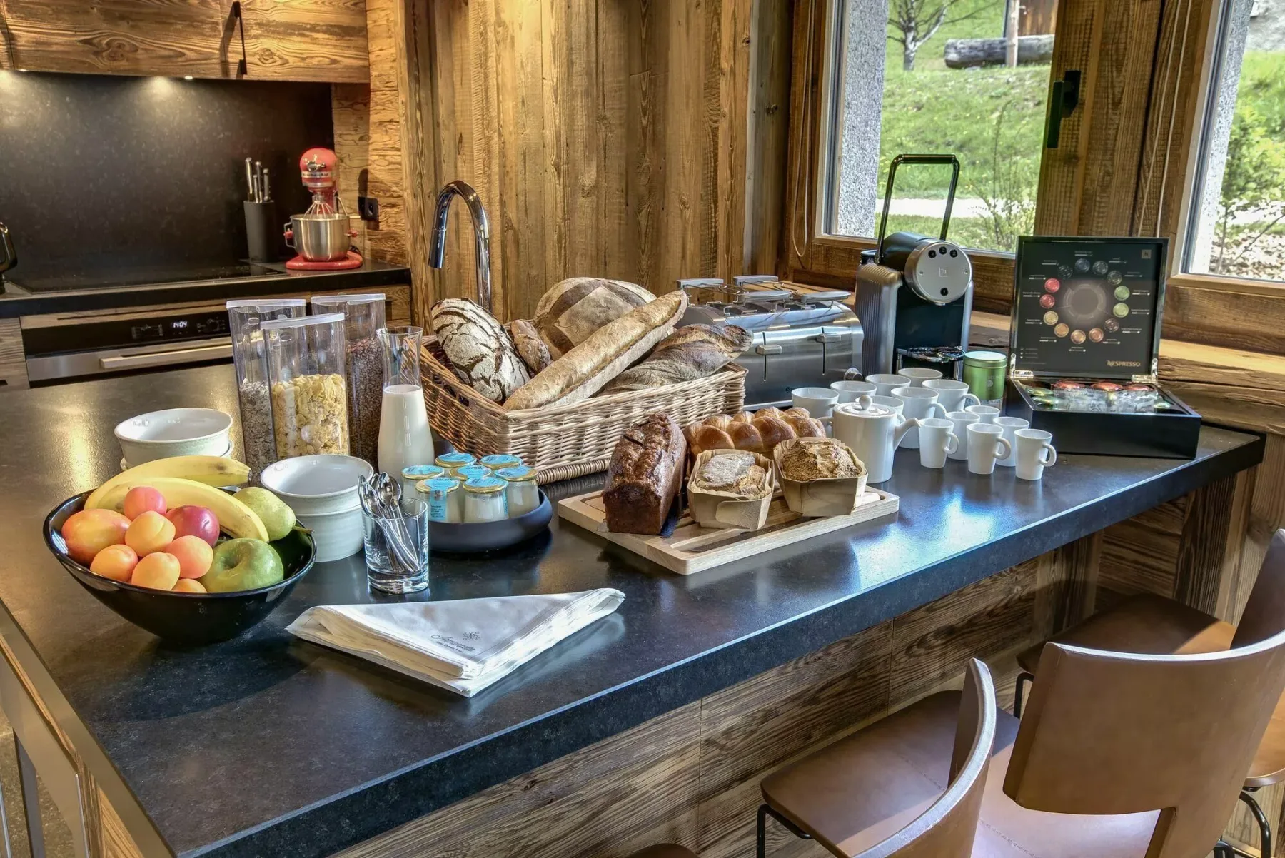 Un bufé de desayuno con pan, fruta, cereales y equipo para café sobre una encimera.
