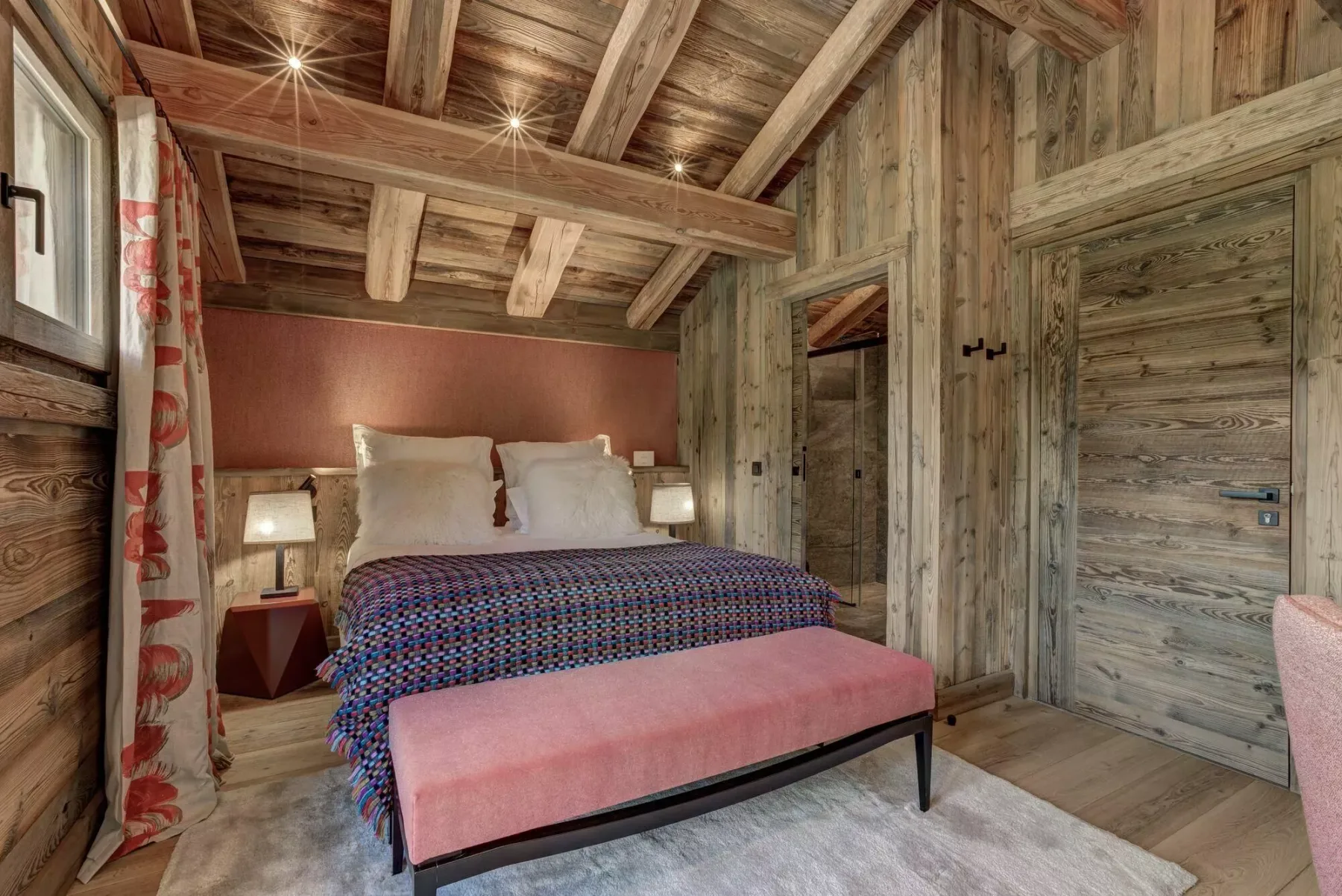 Un dormitorio con una cama, una colcha estampada y un banco rosa bajo un techo de madera.