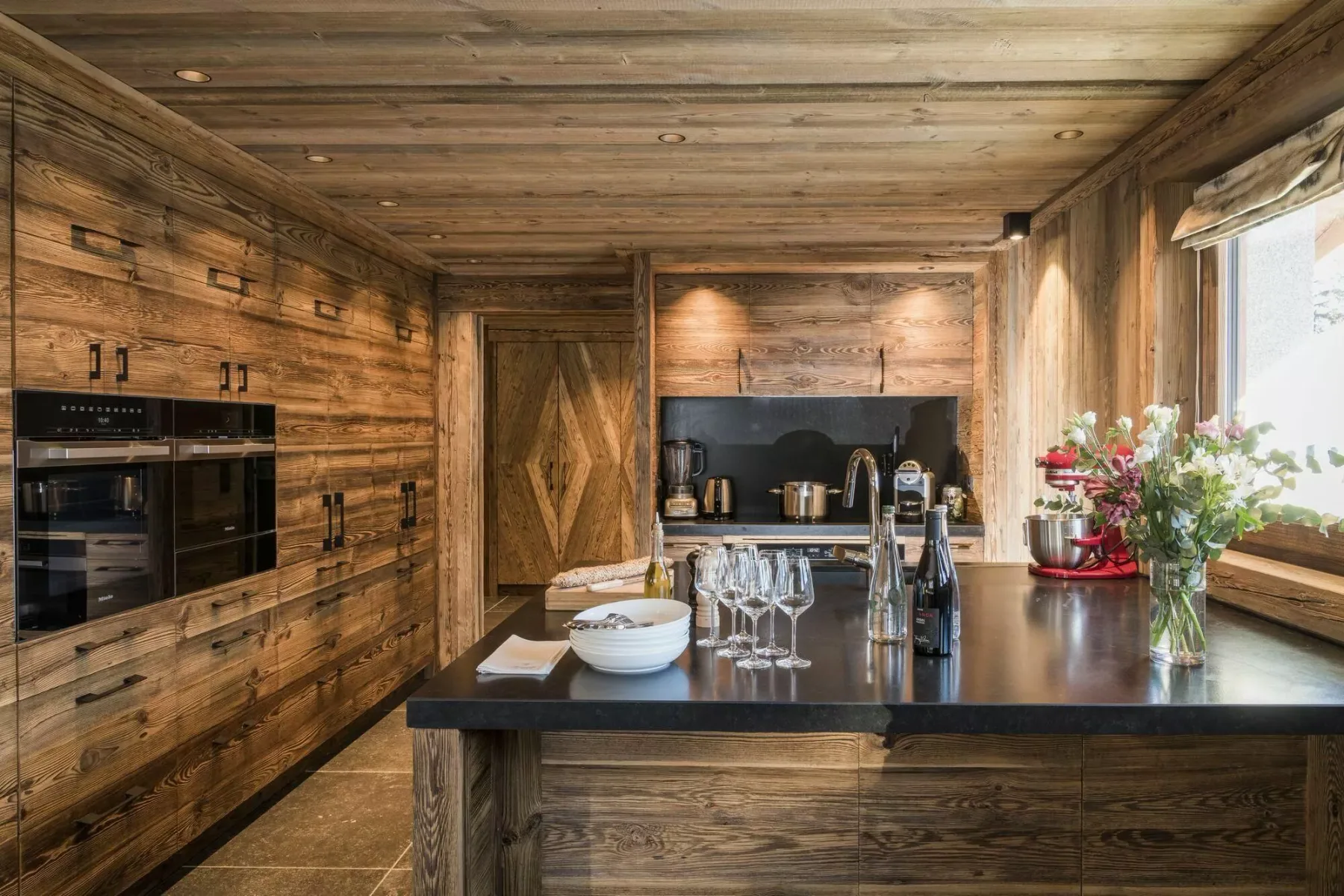 Una cocina revestida de madera tiene una isla central con cristalería y flores junto a la ventana.