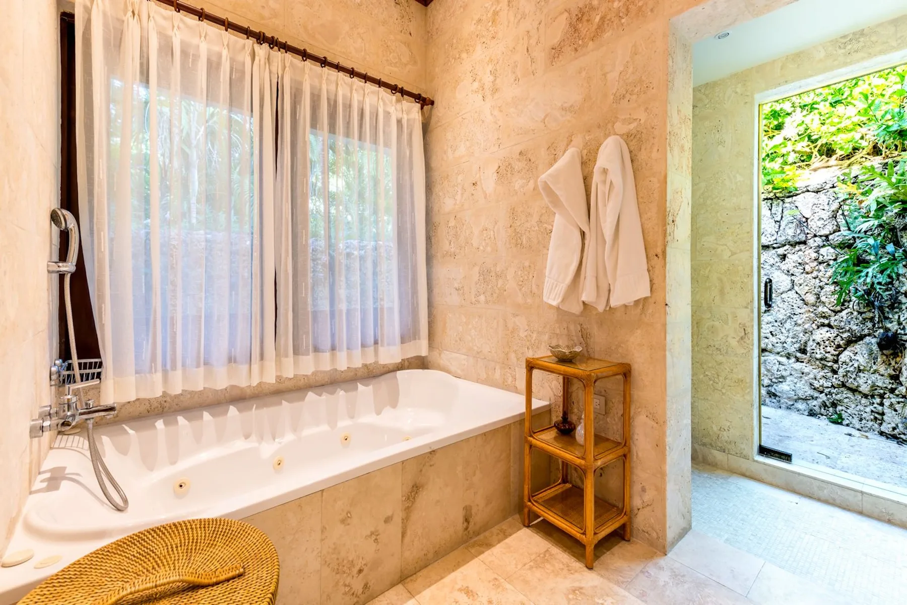 A bright bathroom features a bathtub beside an open doorway to a leafy outdoor area.