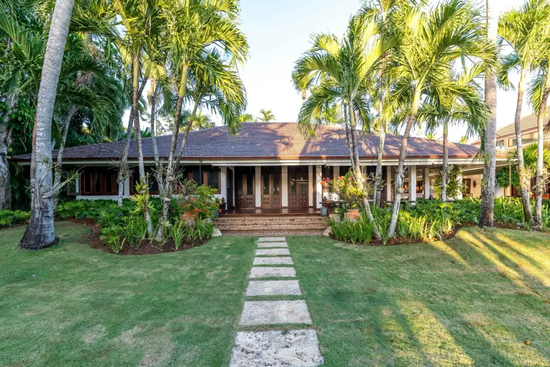 A low building with a covered entrance sits behind a stone path and tropical landscaping.