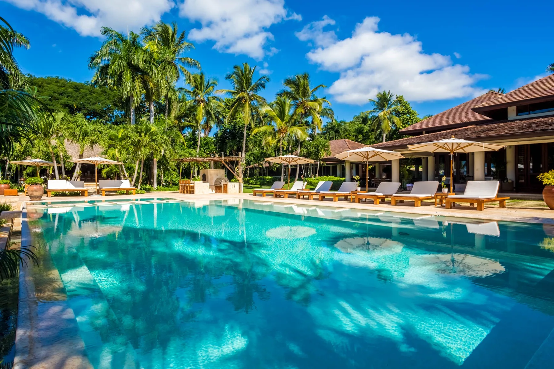 An outdoor swimming pool is surrounded by lounge seating, umbrellas, and palm trees.
