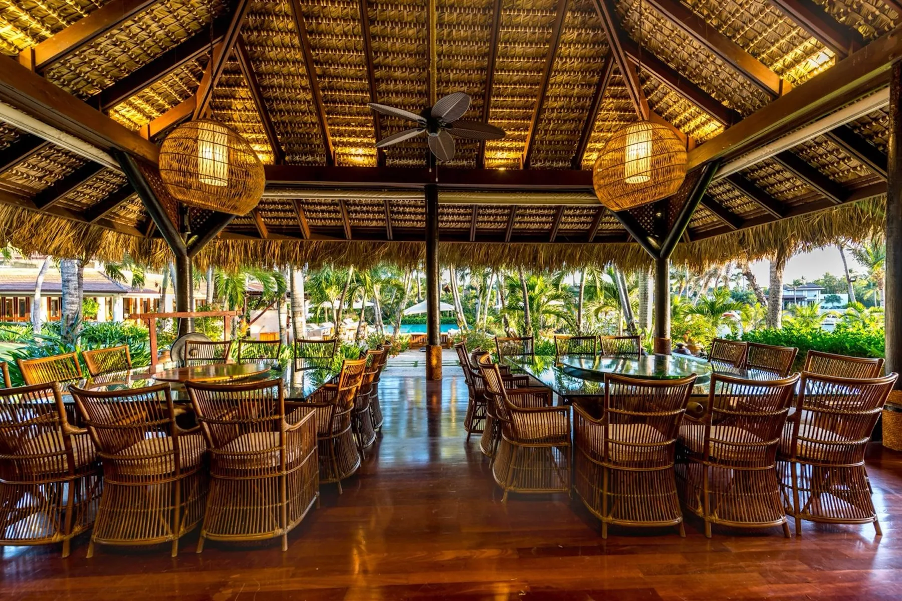 An open-sided dining pavilion with rows of tables and seating beneath a thatched roof.