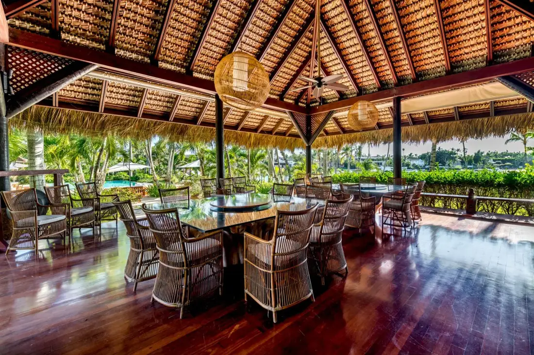 An open-air dining area with tables and seating beneath a thatched roof.