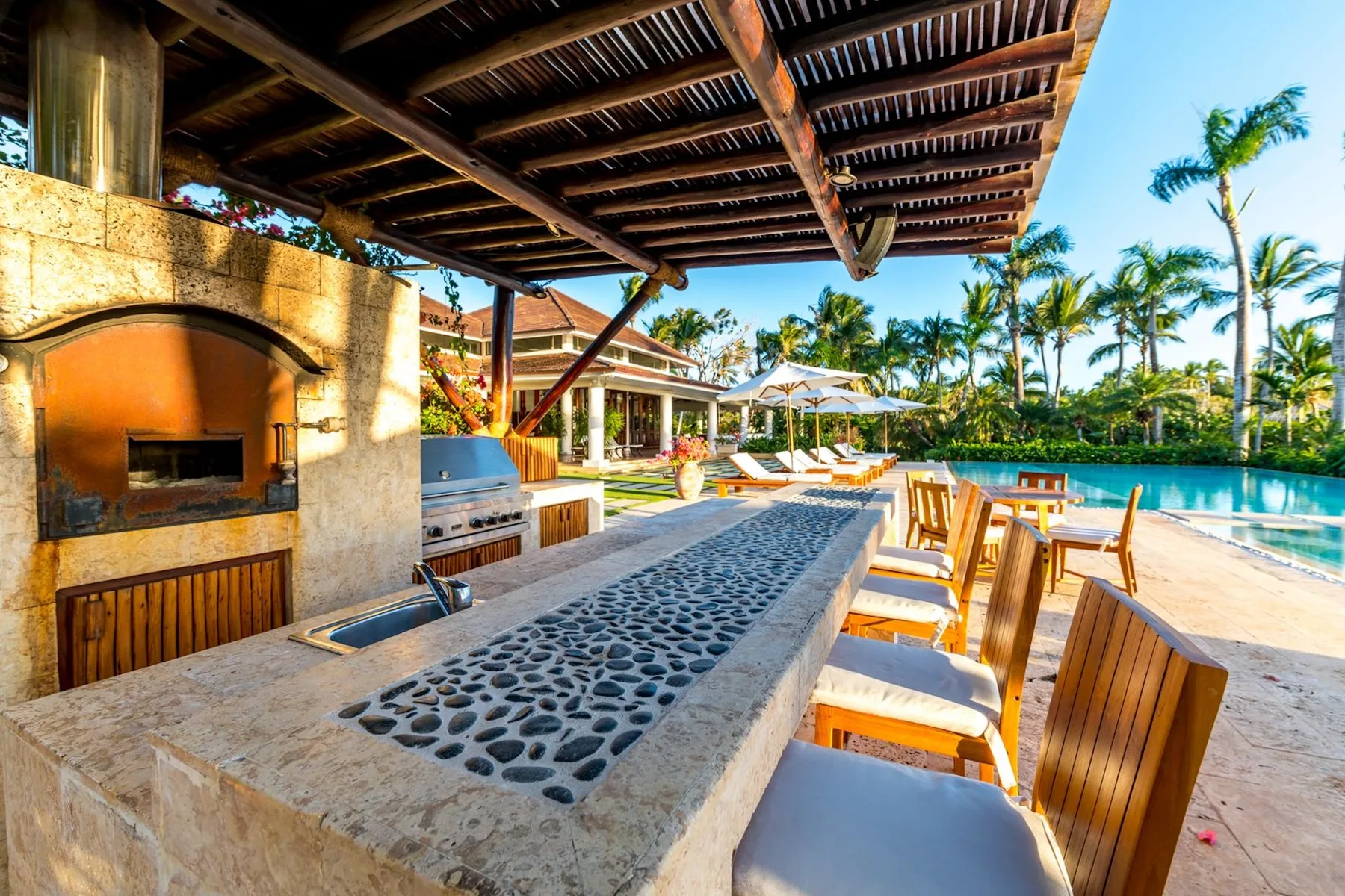 An outdoor kitchen and dining area beside a swimming pool, shaded by a wooden pergola.